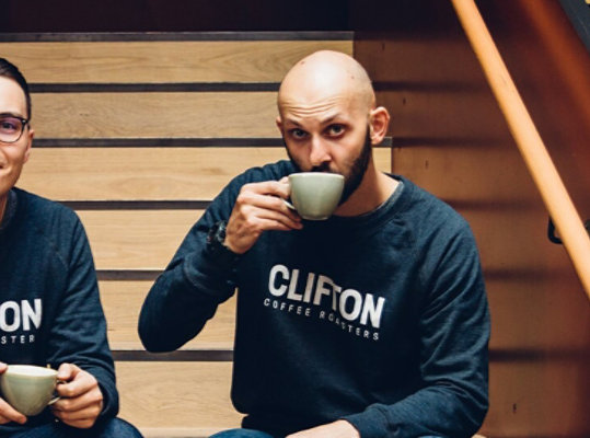 Two people wearing Clinton Coffee Roasters shirts drinking coffee.