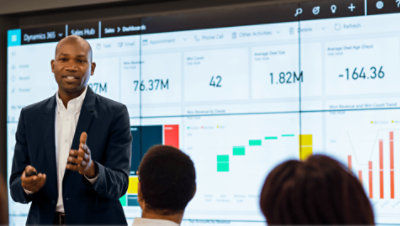 Eine Person, die eine Präsentation hält, während hinter ihr ein Dashboard von Dynamics 365 auf einer Wand von Bildschirmen angezeigt wird