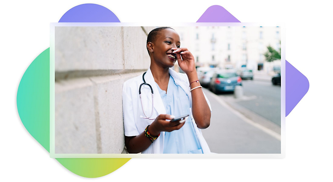 A healthcare worker leaning against a wall holding a mobile phone and laughing.