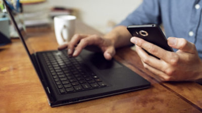 Henkilö kirjoittaa toisella kädellä tietokoneen näppäimistöllä ja pitää toisessa kädessään puhelinta.