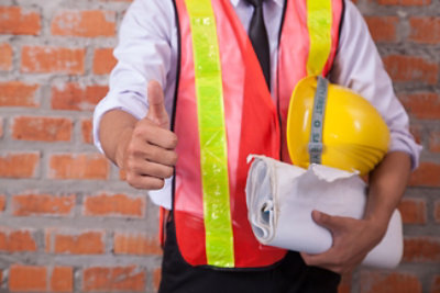 Osoba u prsluku visoke vidljivosti ispred zida od cigala pokazuje podignute palčeve i drži žuti zaštitni šlem i umotane nacrte planova.