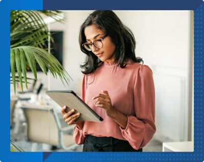 A person wearing glasses stands indoors, holding and looking at a tablet. A plant and an office setting are in the background.