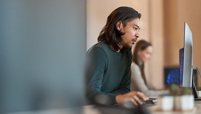 Person working at a desktop computer in an office, with another person blurred in the background