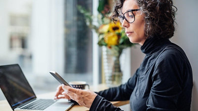 A person working at a laptop while holding a smartphone, seated near a window with a vase of flowers in the background.