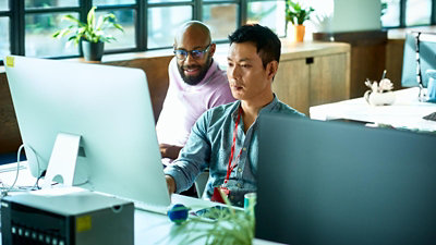 Two people working together at desktop computers in an office, reviewing something on screen.