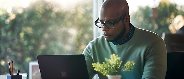 A focused bald man with glasses working intently on a laptop in a bright, plant-decorated office space.