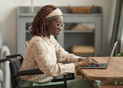 A person in a wheelchair works on a laptop at a wooden table in a well-lit room.
