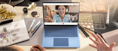 Laptop on a desk with a video call on screen. Desk is cluttered with art supplies, a sketchbook, and small objects.