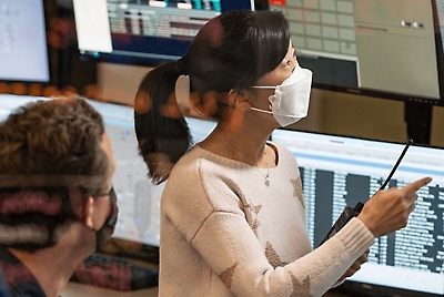 A person wearing a mask holding a walkie talkie and pointing to a screen