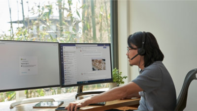 A person wearing a headset works at a desk with two monitors, one displaying a chat application and the other showing a login screen.