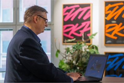 A person in a suit works on a laptop in an office with colorful artwork and plants in the background.