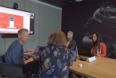 Four people in a conference room engaged in a video call and presentation, with laptops open and a screen displaying slides.