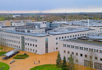 High-angle view of a building with many windows and a curved roof, surrounded by trees and a courtyard with people walking.