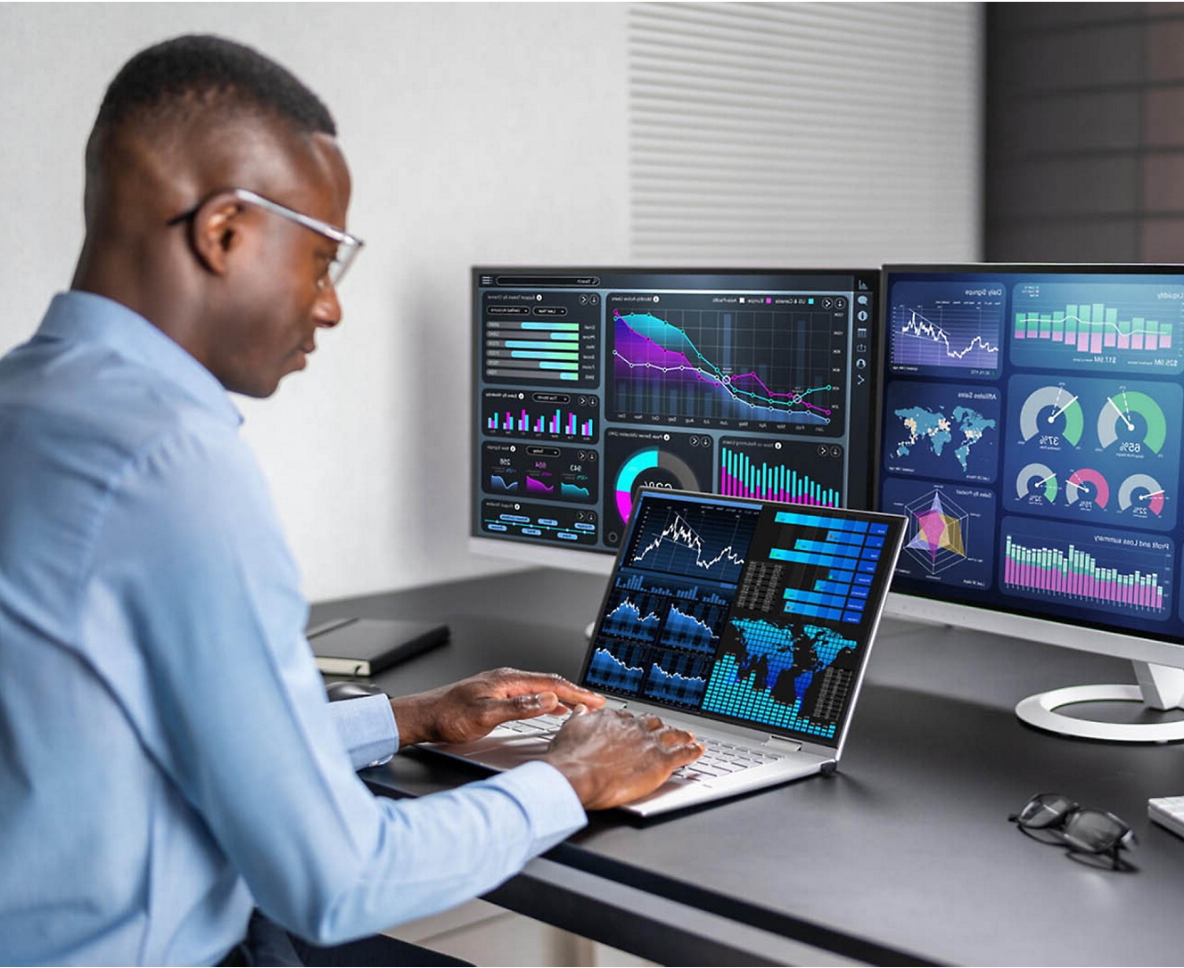 Un hombre con gafas se encuentra en un escritorio, centrado en tres monitores que muestran diversa información.
