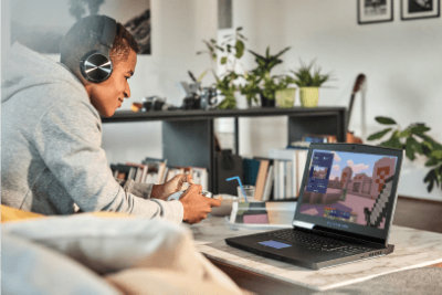 A teenager sitting on the couch, wearing a headset and playing Minecraft on their laptop with a controller.
