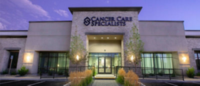 Exterior view of a building with the sign "Cancer Care Specialists" above the entrance, framed by trees and landscaping.
