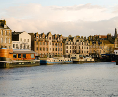 A row of traditional brick buildings and houseboats line the waterfront, under a cloudy sky, reflecting in the calm water.