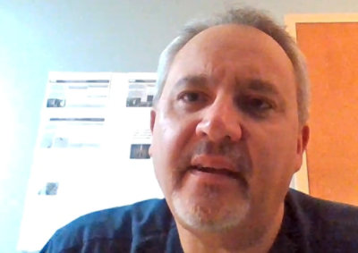 A man with short gray hair and a goatee is speaking. He is indoors, with a blurred background featuring a door and what appears to be a whiteboard or poster.