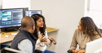 Three people sitting in desk chairs between desks having a conversation
