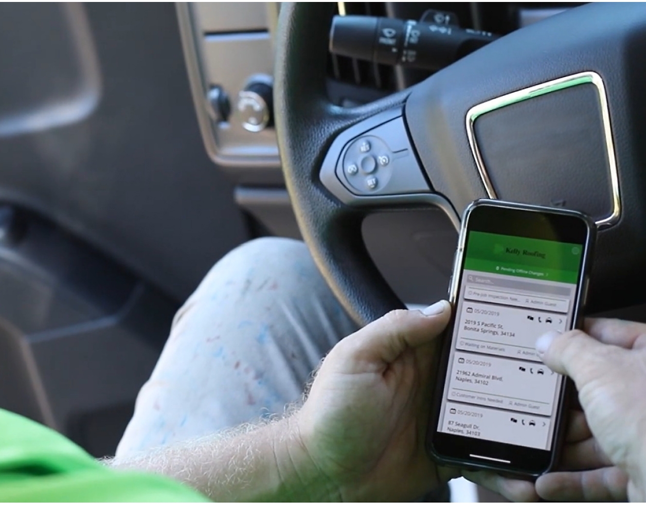 Ein Mann in einem grünen T-Shirt benutzt ein Mobiltelefon, während er einen Lastwagen fährt.