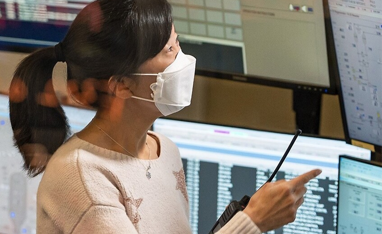 Uma mulher com máscara e um walkie-talkie sentada com vários monitores em sua mesa