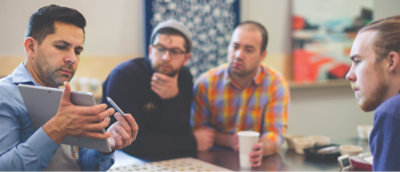 Group of people sitting around a table with one person holding a tablet and another holding a coffee cup.