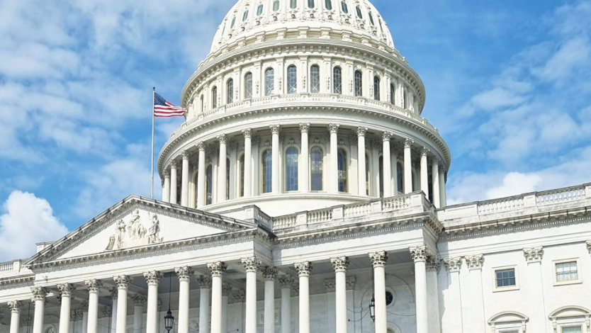 A low-angle, medium shot of the United States Capitol Building in Washington, D.C., showcasing its iconic white dome and neoclassical architecture against a bright blue sky with scattered white clouds. 