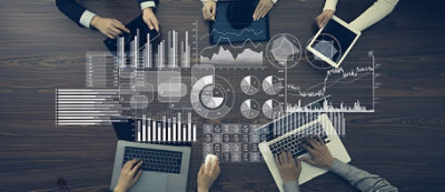 Due mani interagiscono con tablet che mostrano sovrapposizioni di dati, diagrammi e grafici vari su un tavolo di legno.