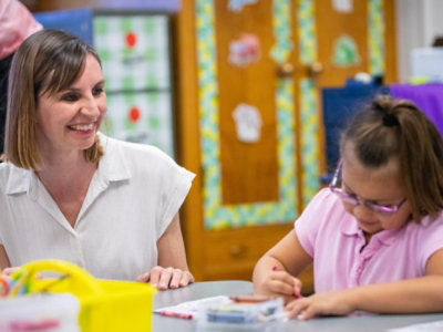 教室のテーブルで絵を描いている若い女の子の隣に座って微笑む女性。