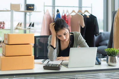 Žena sedi za stolom na kojem se nalaze laptop, kutije, kalkulator i dokumentacija i izgleda zabrinuto. Iza nje