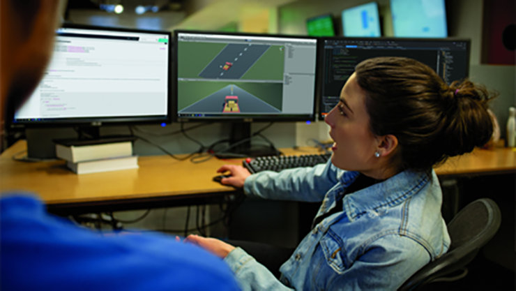 A girl sitting in front of 3 computers and explaining something to another person