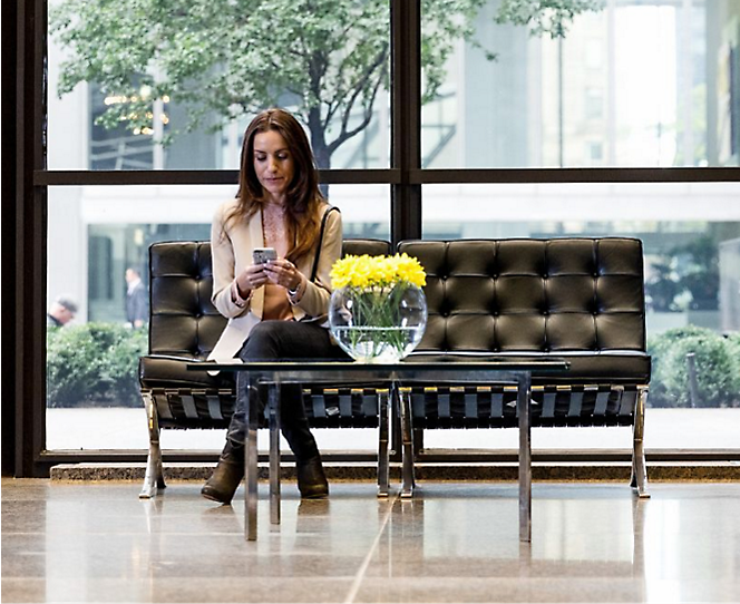 A woman sitting in a waiting area using her phone and on the table a flower vase is present