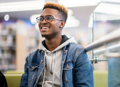 En mann med briller som er iført en denimjakke over en hettegenser, smiler og ser til siden i et rom med sterkt lys.