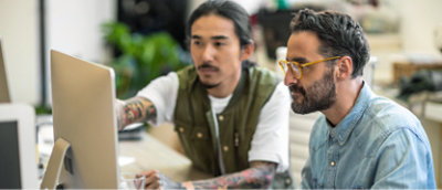 Twee personen die aan een bureau zitten en naar een computerscherm kijken. De persoon aan de linkerkant wijst naar het scherm
