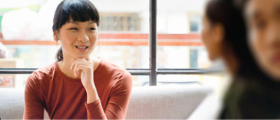 Een persoon met donker haar en een oranje topje zit binnen, lachend en kijkend naar een andere persoon die enigszins wazig is 