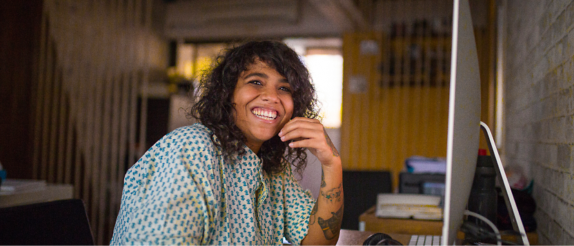 Person with curly hair and tattoos, wearing a patterned shirt, sitting at a desk with a computer, smiling.