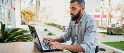 Een man typt op een laptop op een buitenwerkplek, omringd door planten en gebouwen op de achtergrond.
