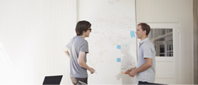 Two men, one in a grey shirt and one in a white shirt, standing in front of a whiteboard.