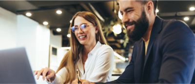 Two people sitting side by side, smiling and looking at a laptop screen in a brightly lit office setting.