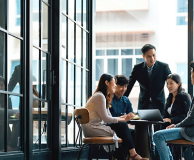 Fem personer som har en diskusjon rundt et bord med en bærbar datamaskin, sitter ved store vinduer i et moderne kontormiljø.