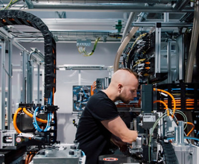 En person arbeider på maskiner i et industrimiljø, omgitt av kabler og teknisk utstyr.