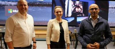 Three people are standing and smiling in front of a large screen displaying various images, including a Microsoft logo. 
