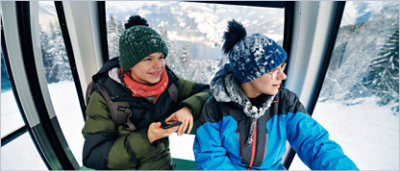 Two people dressed in winter gear sit inside a cable car, surrounded by snowy mountain scenery visible through the windows.