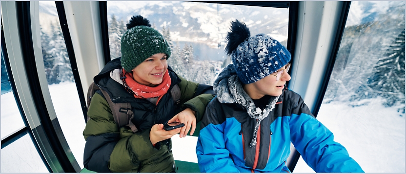 Two people dressed in winter gear sit inside a cable car, surrounded by snowy mountain scenery visible through the windows.