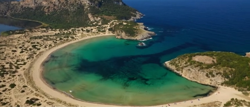 Aerial view of a crescent-shaped beach with clear turquoise water, surrounded by rocky and green landscapes.