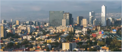 Aerial view of a cityscape featuring numerous buildings and skyscrapers under a cloudy sky, with mountains in the distant background.