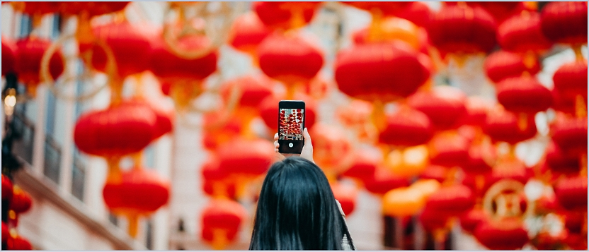 A person holds up a smartphone against a backdrop of numerous red lanterns hanging above a street.