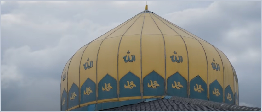 A large, yellow dome with Arabic script, featuring the name of Allah in blue and yellow, is shown against a cloudy sky.