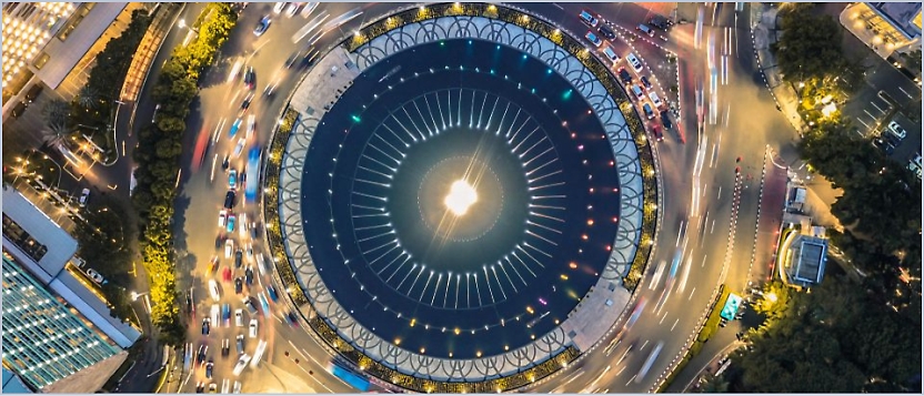 Aerial view of a large, brightly lit circular structure surrounded by busy roads and moving traffic.