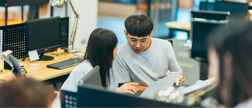 Dos personas hablando sobre un documento en un escritorio de ordenador en una oficina ajetreada.